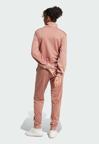Støvet rosa treningsdrakt med lange ermer og høy krage, med ribbede mansjetter og linning. Modellen har på seg hvite joggesko.