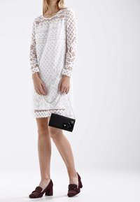 Vestido de encaje blanco con mangas largas que presenta intrincados patrones de diamantes y flores. Combinado con zapatos de tacón burdeos y un pequeño bolso negro.