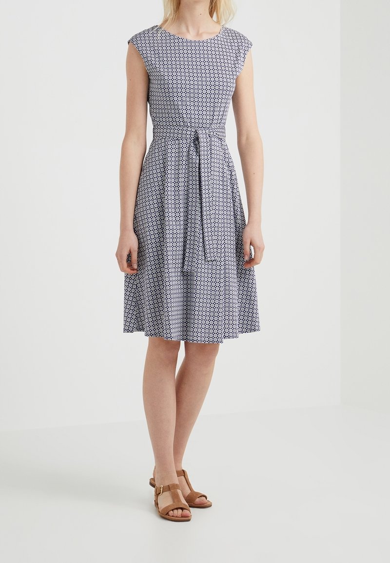 Femme portant une robe sans manches à motifs, longueur genou, avec une ceinture à la taille et des sandales ouvertes marron, debout devant un fond uni.