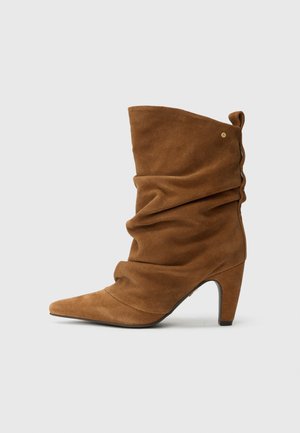 Bottine en daim marron avec un design à effet froissé, un bout pointu et un talon de hauteur moyenne. Elle présente un accent décoratif doré à la cheville.