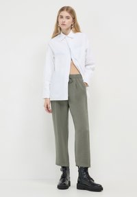 Chemise blanche à boutons, coupe ample, pantalon vert avec taille élastique, bottes de combat noires à lacets avec semelles épaisses, design minimaliste.
