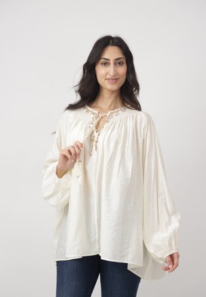Kobieta o długich ciemnych włosach, ubrana w luźną białą bluzkę z haftowanym dekoltem i niebieskie jeansy, stojąca na tle jasnego, jednolitego tła.