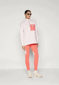 Plyšový pulóver vo svetloružovej farbe s koralovou vreckom, skombinovaný s koralovými legínami s bielymi akcentmi, nosený s svetlými teniskami a slnečnými okuliarmi.