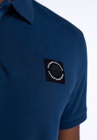 Námorně modrá polo tričko s texturovanou látkou, špičatým límcem a černým čtvercovým nášivkem s kruhovým grafickým detailem.