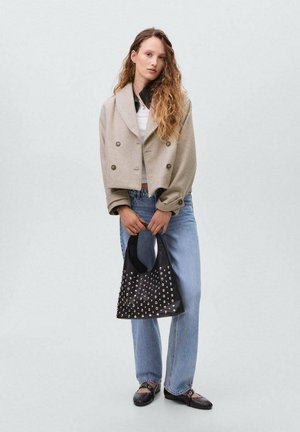 Veste cropped beige à double boutonnage, chemise blanche, jeans évasés bleu clair, sac à main noir à clous, et chaussures noires. Cheveux lisses, arrière-plan neutre.