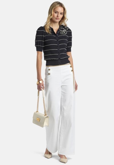 Femeie purtând un top negru cu mâneci scurte și nasturi, cu volane, pantaloni albi cu talie înaltă și croială largă, pantofi cu toc bej și ținând o geantă bej cu lanț auriu.