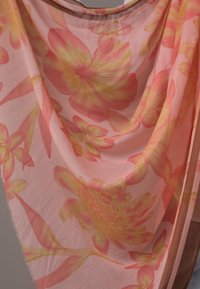 Kevyt kukkahuivi, jossa on suurikokoisia vaaleanpunaisia ja keltaisia kukkia pehmeällä vaaleanpunaisella taustalla, reunat hieman rispaantuneet ja huivi laskeutuu sulavasti.