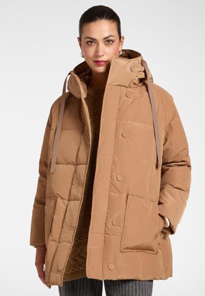 Frau trägt eine beige gesteppte Kapuzenjacke mit großen Fronttaschen und Knopfverschluss, kombiniert mit einem braunen Pullover und grauer Hose.
