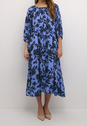 Frau trägt ein blaues Midikleid mit dunklem Blumenmuster und 3/4-ärmeln mit Volants, kombiniert mit beigen spitzen Pumps.