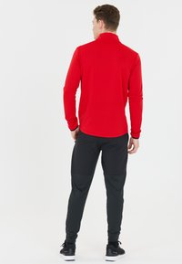 Rode sportieve top met lange mouwen en hoge kraag, gecombineerd met zwarte taps toelopende broek met textuur- en gladde secties, gedragen met zwarte sneakers.
