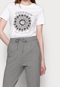 Hvid t-shirt med sort mandala print og teksten "FRIHED" samt "STORME VARER IKKE EVIGT"; parret med grå sweatpants med elastisk snoreliv.