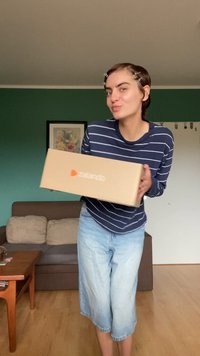 Caixa de cartão da Zalando em castanho claro com um logótipo laranja, segurada por uma pessoa vestindo uma camisa de mangas compridas às riscas em azul-marinho e branco e calções azuis claros.