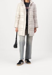 Osoba nosící béžový prošívaný kabát, světle modré džíny, černé tenisky s bílými pruhy, držící malou černou kabelku.