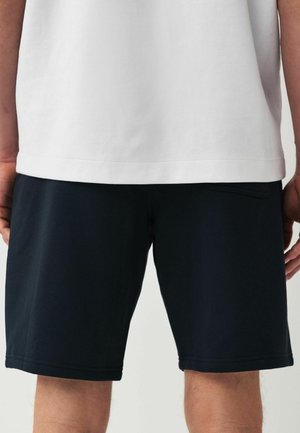 Personne portant un short noir et une chemise blanche, vue de la taille vers le bas, se tenant contre un fond uni.