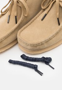 Beige velúrbakancs krém talppal, barna fűzőkkel és fekete csúszkákkal, két sötétkék fűzőpótlékkal együtt.