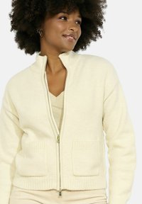 Kremni zip-up cardigan iz mehke tkanine, z rebrastimi detajli, dvema sprednjima žepoma in visokim ovratnikom.