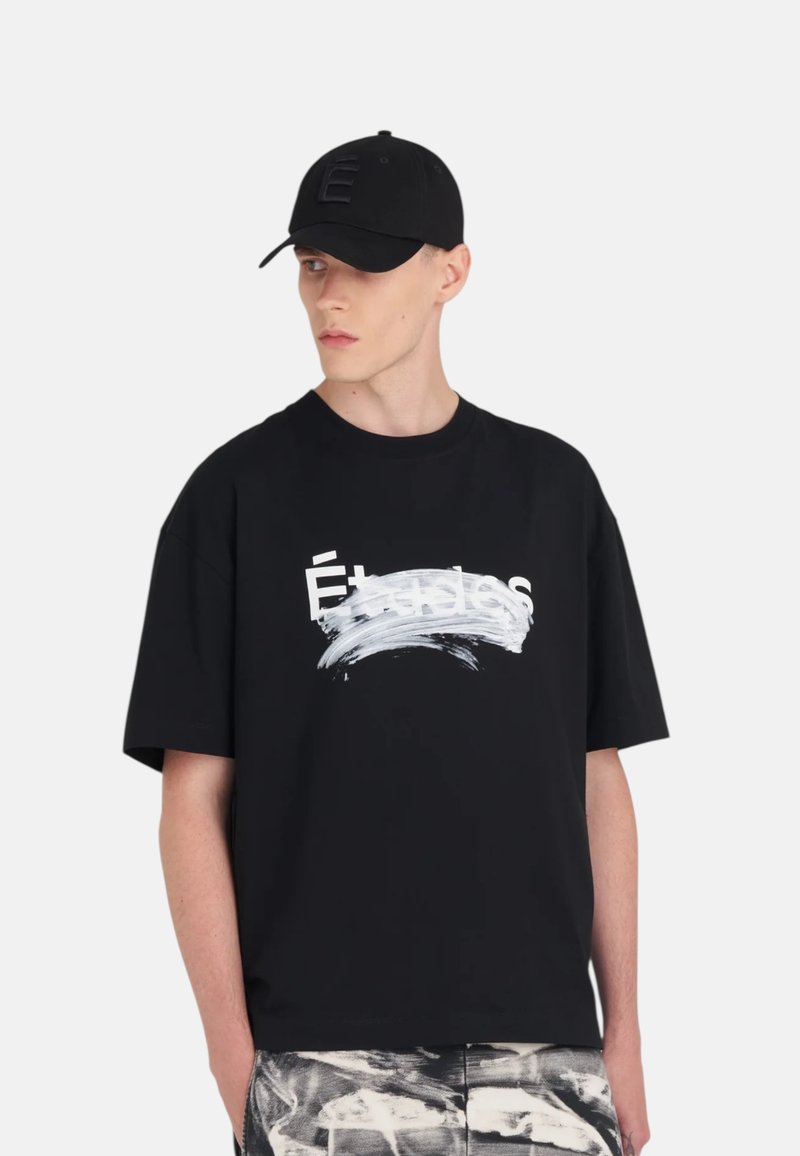 T-shirt noir à manches courtes avec un motif de coups de pinceau blancs et du texte imprimé. Porté avec une casquette noire portant un logo "E" texturé.