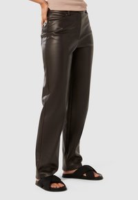 Pantalon en cuir synthétique marron avec une coupe droite, équipé de poches latérales et d'une texture lisse. Associé à des sandales noires à enfiler.