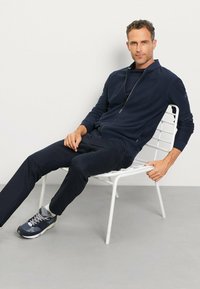 Marinefarbener Reißverschluss-Hoodie und passende Hose aus weichem Stoff. Das Model sitzt auf einem weißen Stuhl und trägt graue und marineblaue Sportschuhe.