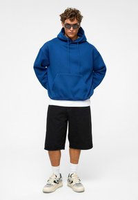 Un sweat à capuche bleu avec des cordons de serrage et une poche avant, associé à un short noir, un t-shirt blanc et des baskets avec un design texturé.