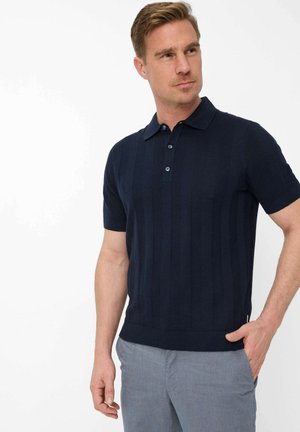 Mann trägt ein marineblaues Kurzarm-Poloshirt und hellgraue Hose, steht mit einer Hand in der Tasche und blickt seitlich vor einem schlichten Hintergrund.