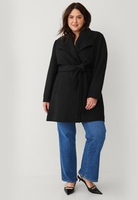 Sort indpakningsfrakke med stor krave og bælte, der har to forlommer, parret med blå lige ben jeans og spidse sorte sko.