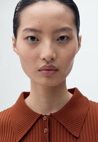 Gros plan sur une personne portant une chemise côtelée de couleur rouille avec un col à boutons. Peau lisse avec des traits subtils, arrière-plan neutre.