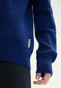 Donkerblauwe trui met een gestructureerd rasterpatroon en geribbelde manchetten. Beschikt over een klein label aan de zijkant. Gemaakt van een gebreide stof.