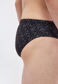 Nærbilde av en person iført svarte badebukser med gjentatt hvit "ARENA" tekstmønster på lys bakgrunn.