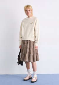 Krämfärgad sweatshirt med broderad logga, ihopparad med en lager-på-lager brun rutmönstrad kjol. Modellen har på sig rosa snörskor och vita strumpor.