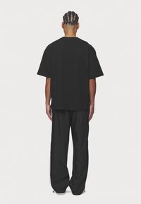 Osoba z warkoczami stoi tyłem, ubrana w luźną czarną koszulkę i czarne spodnie z białymi paskami po bokach na jednolitym tle.