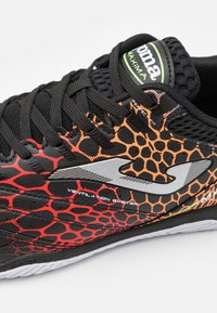 Zapatilla deportiva negra con un upper texturizado que presenta un patrón de panal en rojo y naranja, cordones reforzados y un acento plateado en el logo.