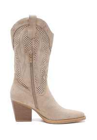 Beżowy zamszowy kowbojski but w stylu Western, z spiczastym czubkiem, ozdobnymi wycięciami oraz bocznym zamkiem. Posiada trzewik na słupku i wytłaczane detale.