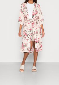 Kwiatowy kimono w kolorze jasnoróżowym, z luźnymi rękawami, paskiem do przewiązania oraz intensywnymi wzorami róż i zieleni. W zestawie z beżowymi klapkami.