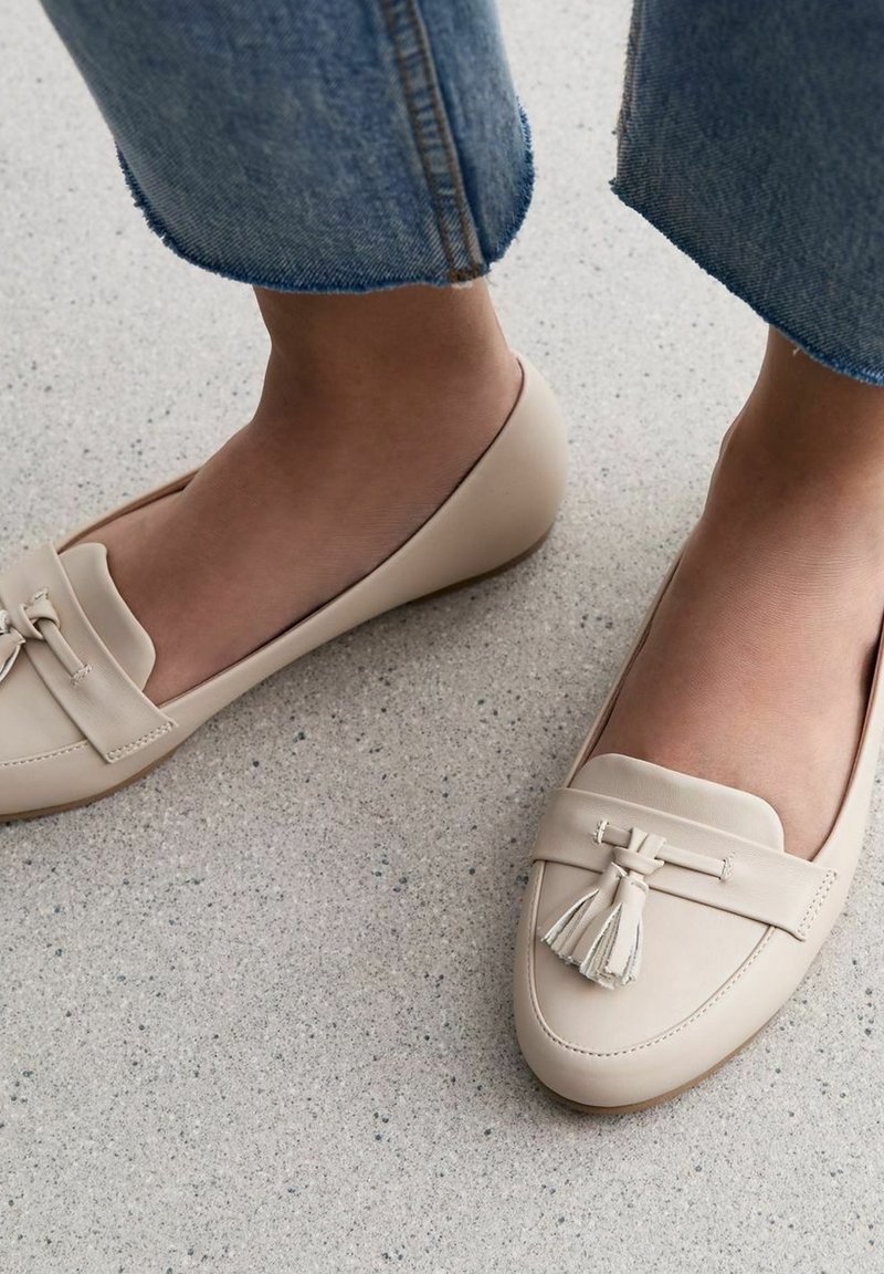 Mocassins en cuir beige avec un accent à frange, bout arrondi et semelle plate, associés à un jean en denim bleu retroussé. Texture lisse.