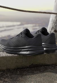 Paire de chaussures de course Adidas noires avec semelles Cloudfoam rembourrées reposant sur un rebord en pierre à l'extérieur près d'un tronc d'arbre.