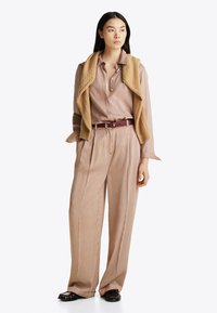 Beżowa koszula w paski z guzikami, szerokie spodnie, a na ramionach jasnobrązowy sweter w warkocze, zabezpieczony ciemnym paskiem.