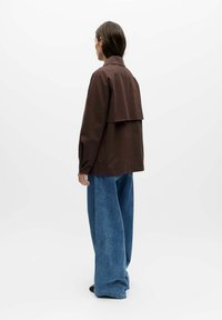 Femme portant une veste marron avec un rabat dans le dos et un jean large bleu, debout de dos avec la tête tournée sur le côté sur un fond blanc.
