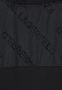 Schwarzes Oberteil mit gestepptem Design und ausgestanzten Buchstaben, strukturiertem Stoff und glattem unteren Abschnitt. Horizontale Nahtdetails sind sichtbar.