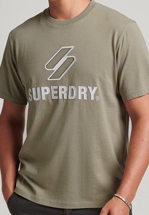 Person trägt ein olivgrünes T-Shirt mit weißem Superdry-Logo und geometrischem Design auf der Brust, Hände ruhen nahe den Taschen.