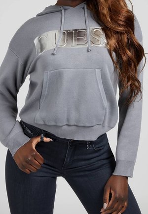 Woman wearing a grey cropped hoodie with metallic "GUESS" logo, paired with dark high-waisted jeans, posing with one hand on hip.