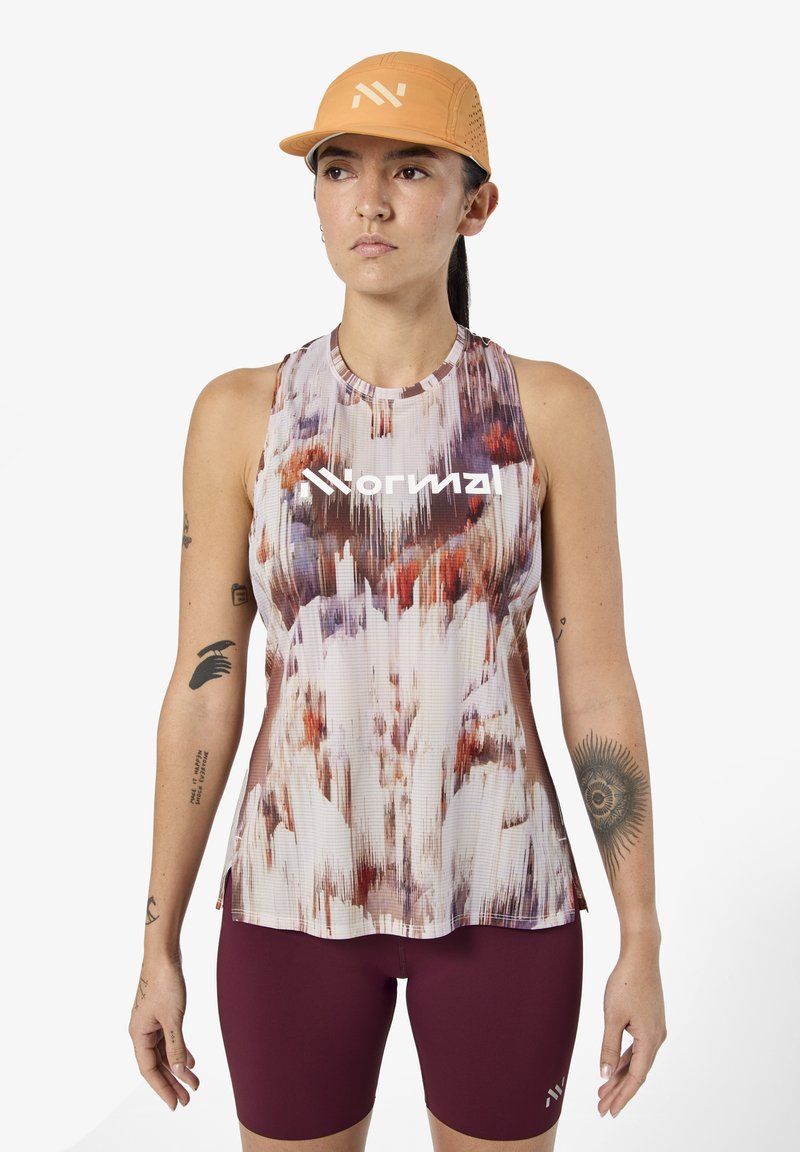 Γυναίκα που φοράει αμάνικο πάνω μέρος προπόνησης με μοτίβο και λογότυπο "normal", μπορντό ποδηλατικές βερμούδες και πορτοκαλί καπέλο, στέκεται μπροστά σε λευκό φόντο.