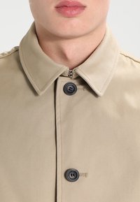 Cappotto beige con chiusura a bottoni e colletto classico, dotato di bottoni neri e una texture di tessuto liscia e leggermente lucida. Dettaglio con zip sul collo.