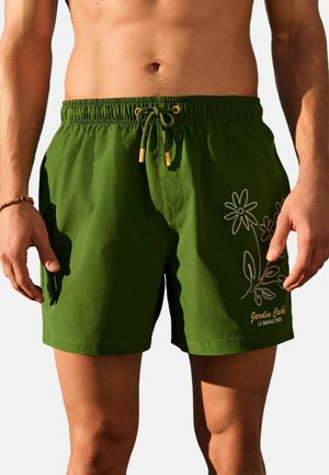 Man wearing green swim shorts with floral embroidery and text "Jardin Caché Le Marais, Paris" on the right leg, standing against a light background.