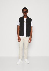 Gilet nero senza maniche con zip frontale, indossato sopra una t-shirt bianca con testo dorato, abbinato a pantaloni beige e sneakers bianche.