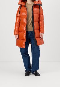 Puffer coat in bright orange with a shiny finish, featuring a hood, black accents, and a zip closure, paired with blue relaxed-fit jeans.