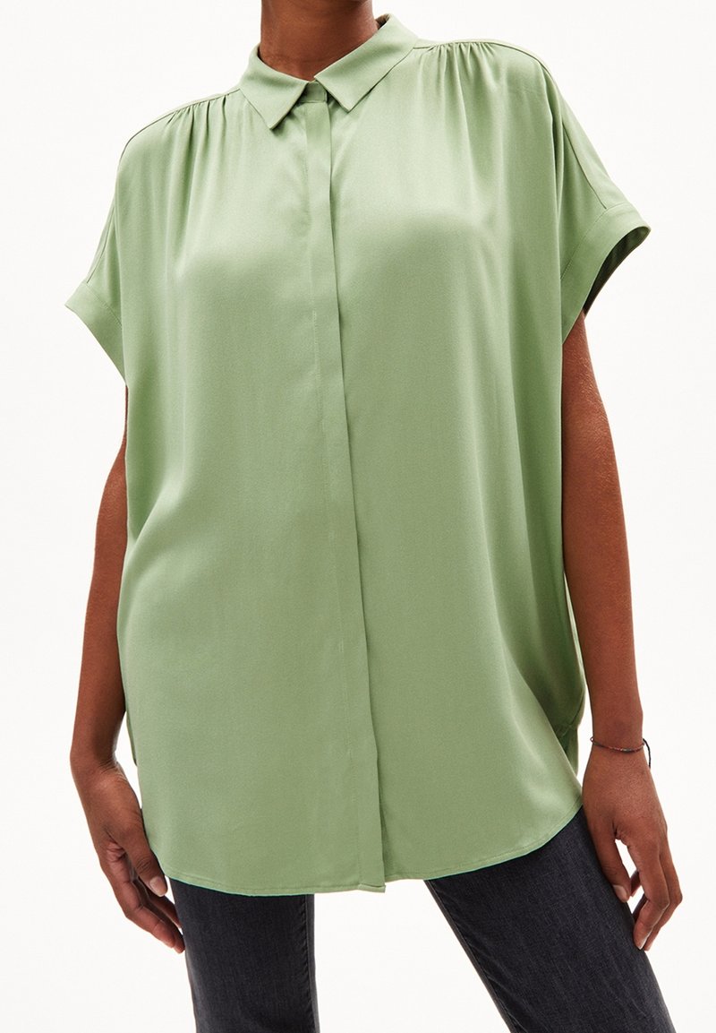 Femme portant un chemisier ample vert sauge à manches courtes avec des boutons cachés à l'avant et un col large, assorti à un pantalon foncé.