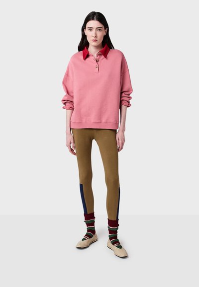 Kvinna iklädd en rosa tröja med röd krage, bruna leggings med marinblå sidremsor, randiga strumpor och beige skor, stående mot en neutral bakgrund.