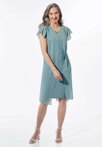 Een licht turquoise jurk met fluistermouwen, een sleutelgat-halslijn en een rechte belijning, gemaakt van zachte, vloeiende stof. Bevat zijsplitten.