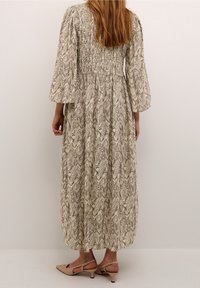 Femme se tenant dans une robe crème à manches longues et longueur jusqu'aux chevilles avec un motif marbré marron, portant des chaussures à talons bas beiges avec un dos ouvert.